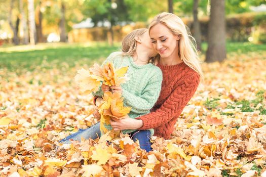 Happy young caucasian mother and little daughter holding autumn yellow leaves sitting and kissing at the park