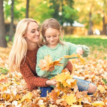 Happy young caucasian mother and little daughter holding autumn yellow leaves sitting at the park