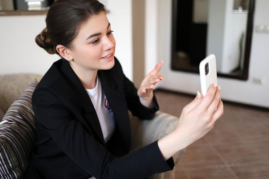 Attractive smiling young woman in a black jacket talking on a mobile video link while sitting in a chair, gesturing with her hand