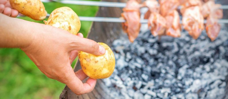 Hands of man prepares barbecue meat with potatoes on skewer by grill on fire outdoors. Concept of lifestyle rustic food preparation