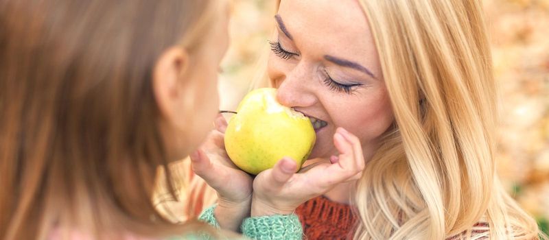 Portrait of little girl with young mother eating yellow apple in autumn park