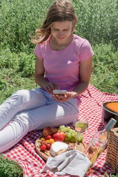 woman in white pants and pink shirt outside having picnic and using smartphone taking photo. Summer fun and leisure