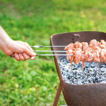 Hands of man prepares barbecue meat on skewer by grill on fire outdoors