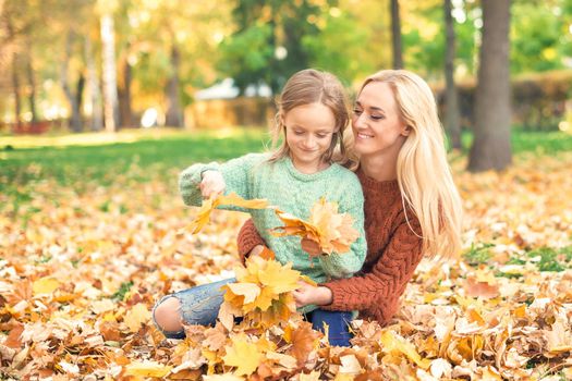 Happy young caucasian mother and little daughter holding autumn yellow leaves sitting at the park