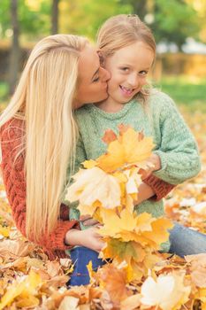 Happy young caucasian mother and little daughter holding autumn yellow leaves sitting and kissing at the park