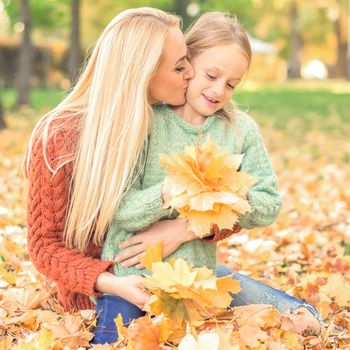 Happy young caucasian mother and little daughter holding autumn yellow leaves sitting and kissing at the park