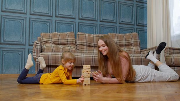 Happy young mother woman teaching small child daughter playing wooden blocks board game at home. Overjoyed little kid girl concentrating learning, enjoying fun hobby time with parent mum or nanny