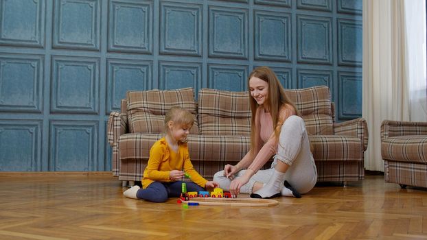 Mother with little daughter child girl riding toy train on wooden railway blocks board game. Family enjoy playtime together sit on floor in living room at home. Kid developmental game, leisure hobbies