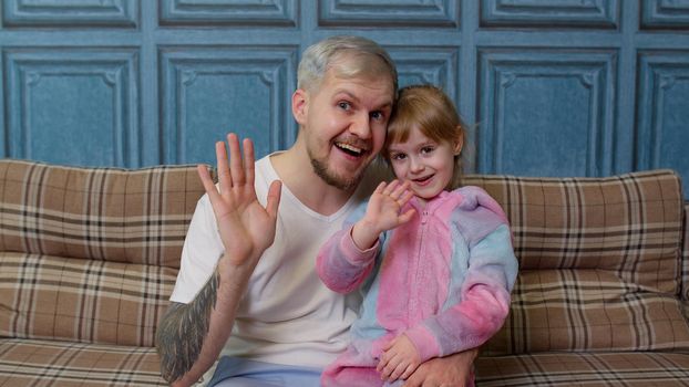 Father and little child daughter kid in pajamas sit on couch in room smiling, looking at camera, joyfully greeting, waving hi, hello, welcome together. Girl with young man. Bonding family generation