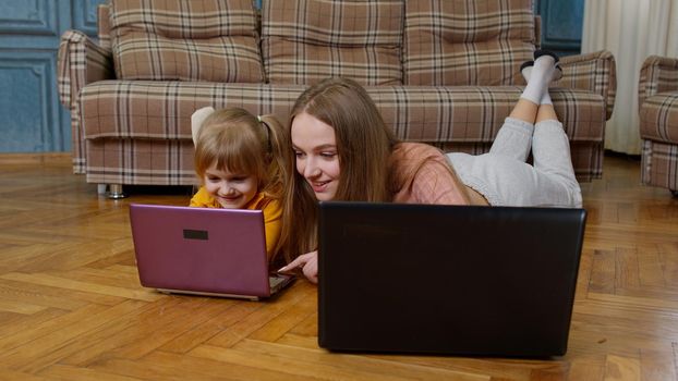 Woman nanny and child girl studying together with computer laptop, while lying on warm floor at home. Family mother and cute kid working, typing on pc, kid watching online education lessons, cartoons