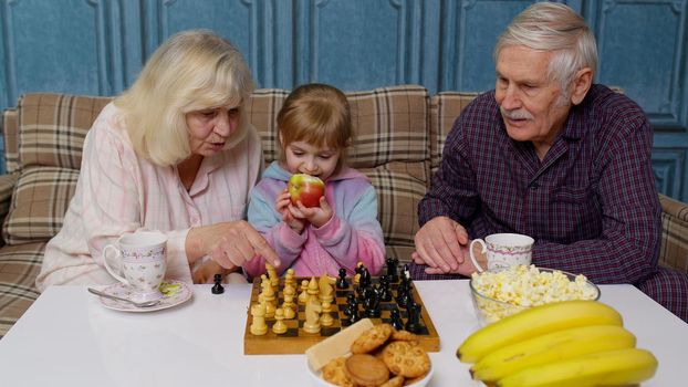 Smiling senior couple grandfather, grandmother resting on sofa, drinking coffee, playing chess with granddaughter child girl kid at home. Retired husband and wife grandparents enjoying play board game