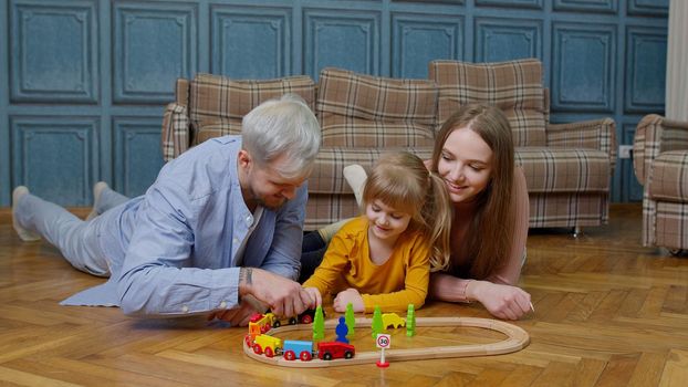 Young mother and father playing with small child daughter riding toy train on wooden railroad blocks board game at home. Little kid girl concentrating learning, enjoying fun hobby with parents family