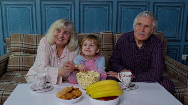 Grandparents watch TV with granddaughter at home. Senior grandfather and grandmother happy laughing, using family time relax with young girl kid sitting on sofa in living room watching cartoons movies