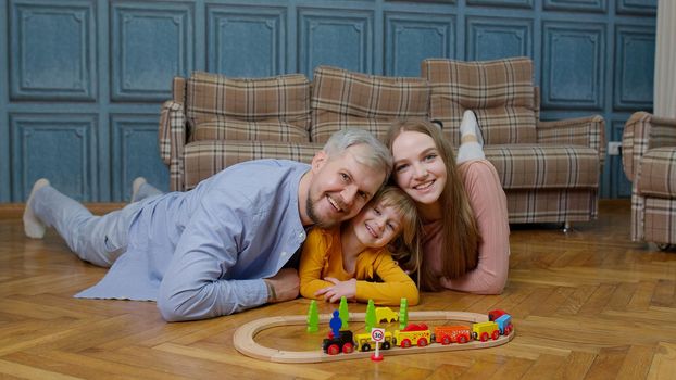 Mother and father with daughter child girl riding toy train on wooden railway blocks board game. Family enjoy playtime together on floor in living room at home. Kid developmental game, leisure hobbies