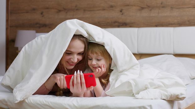 Joyful mother lying on bed under duvet blanket with daughter kid girl, holding cell phone, watching cartoons movies, using funny mobile social media apps, making online shopping together in bedroom