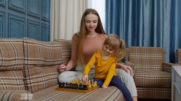 Happy young mother woman teaching small child daughter playing chess on wooden board at home sofa. Overjoyed little kid girl concentrating learning board game enjoying fun hobby time with mum or nanny