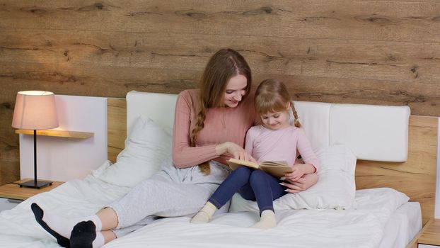 Young mother and teen child daughter lying in bed at home bedroom on duvet blanket and reading book bedtime stories and fairytales together with warm lamp at night. Family bonding, parenthood concept