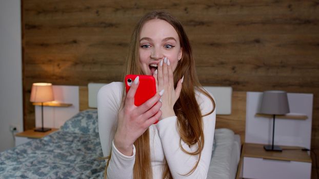 Surprised shocked young woman sitting on bed in bedroom. People lifestyle concept. Say wow put hand on head showing amazed like gesture, holding mobile phone. Perfect exciting news. Impressed reaction