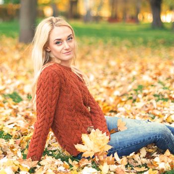 Beautiful young caucasian woman in red sweater sitting on the leaves in the autumn park