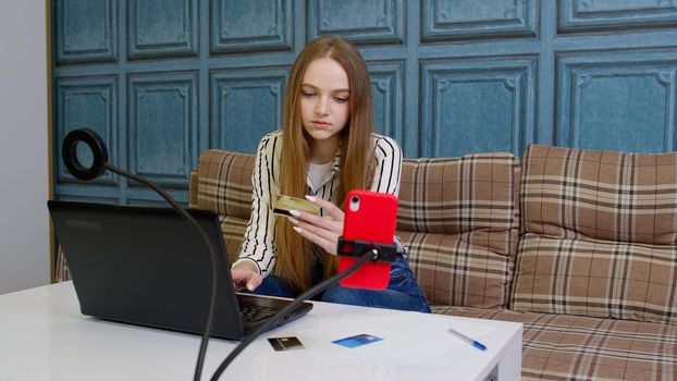 Young woman sitting on couch in modern living room apartment, ordering food, making shopping payments purchases transaction on Internet from bank credit card on laptop. Online banking with computer