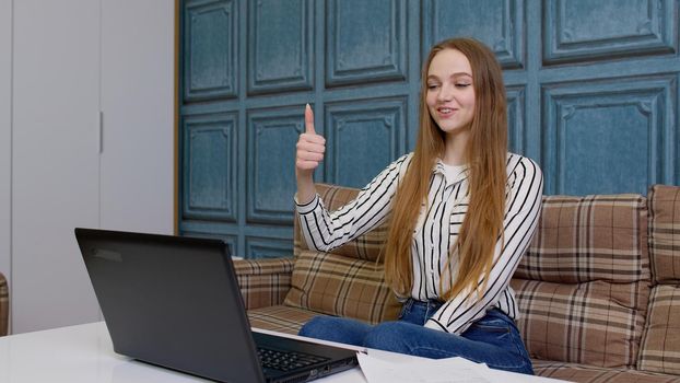 Young business woman sitting at workplace home office, making video call looking at laptop computer screen reads good news shows on camera like puts thumbs up makes gesture of success approval