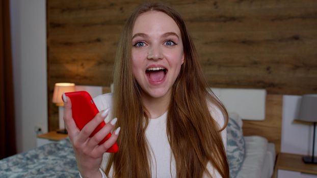Happy young woman customer celebrating winning bid or getting ecommerce shopping discount offer on smartphone. Excited girl winner looking at mobile phone using app celebrating success at home concept