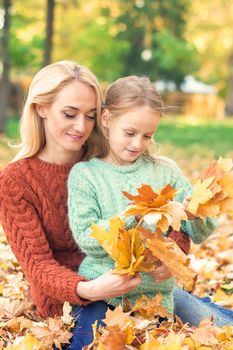 Happy young caucasian woman and little girl holding autumn yellow leaves sitting at the park