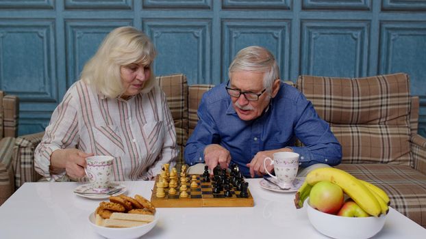 Retired senior couple talking drinking tea, playing chess in modern living room lounge together. Old elderly grandmother, grandfather enjoying coffee having conversation relaxing sit on sofa at home