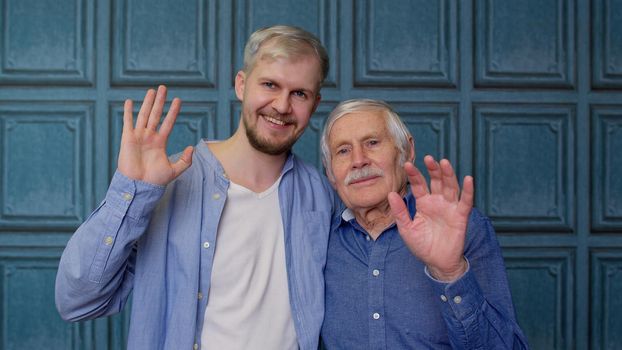 Happy bonding loving different male generations family of senior father and handsome adult son or grandson waving hi, hello, welcome. Cheerful excited young man with gray-haired old dad or grandfather