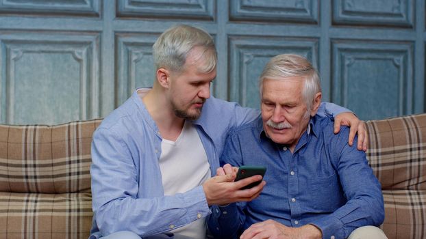 Happy different male generations family senior father and son watching on online mobile phone internet with fun, elderly and young man leisure at home relax together, resting, communication, lifestyle