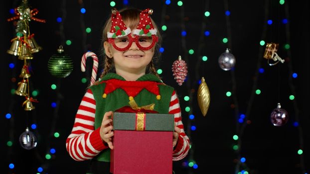 Lovely teen kid girl in Christmas elf Santa helper costume isolated on black background with garland. Child giving present gift box to camera, smiling sweetly. People New Year holidays celebration
