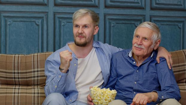 Senior and young men watching football match on television. Caucasian different males generations family. Pensioner old father and adult son sitting on sofa at home and cheering up for goal or victory