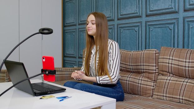 Smiling young business woman professional blogger talking, recording vlog on mobile phone, using laptop sit at home office, customer make video call confirming online website shopping order delivery
