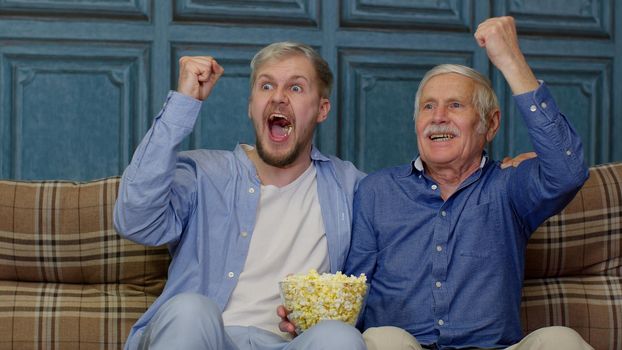 Caucasian different males generations family. Pensioner old father and adult son sitting on sofa at home and cheering up for goal or victory. Senior and young men watching football match on television