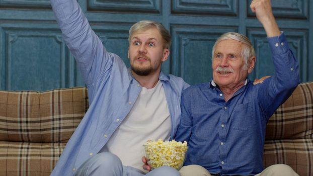 Happy different male generations family watching TV, shouting, celebrating emotionally football championship win at home. Overjoyed senior mature man supporting favorite sport team with grown up son.