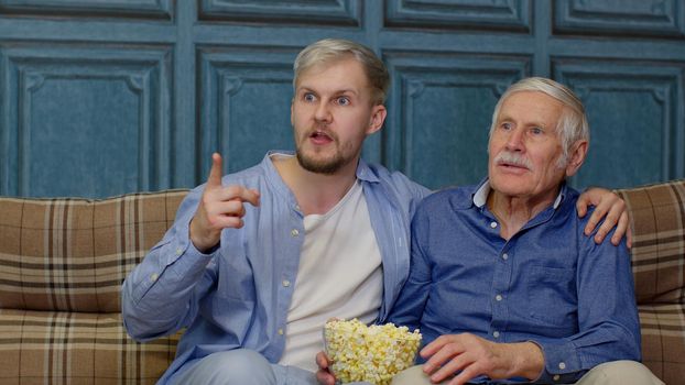 Dad and son cheering soccer or football team to winning the match. Excited Caucasian family senior man father and adult son sitting on sofa and watching sport game on TV with eating popcorn together.