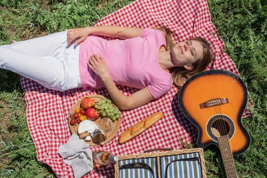 Top view of young caucasian woman in white pants outside having picnic, eating and playing guitar. Summer fun and leisure