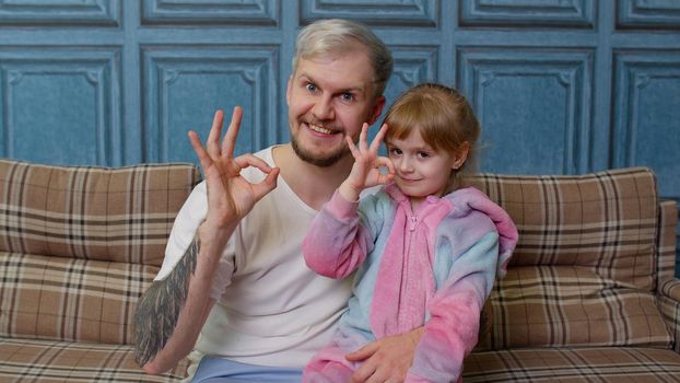 Father and little child daughter kid in pajamas sit on couch in living room smiling, looking at camera, embracing, showing ok gesture sign together. Girl with young man. Bonding family generation