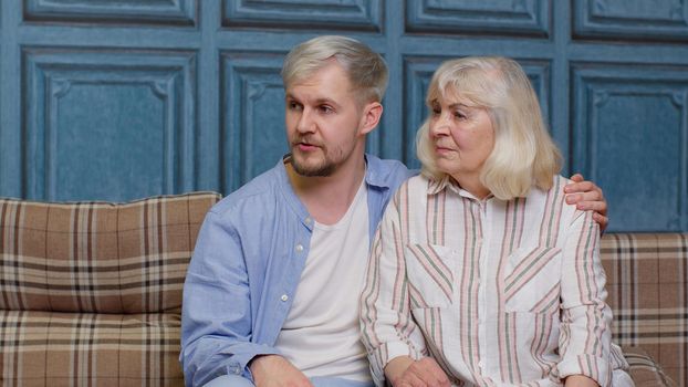Cheerful generations family of senior mother embracing adult son or grandson and having heart-to-heart conversation. Young man hugging gray-haired old mom or grandmother giving good psychological help