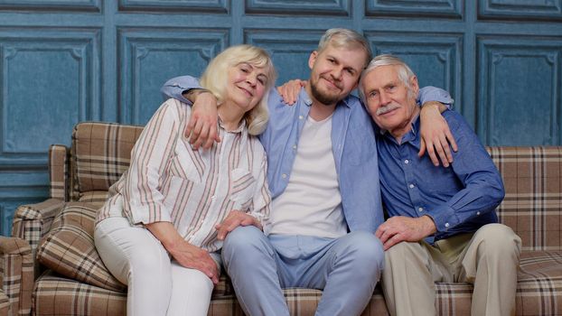 Portrait of happy generation family of senior grandparents with adult son embracing having fun looking at camera bonding at home, loving mother father. Man hugging grandmother and grandfather on sofa