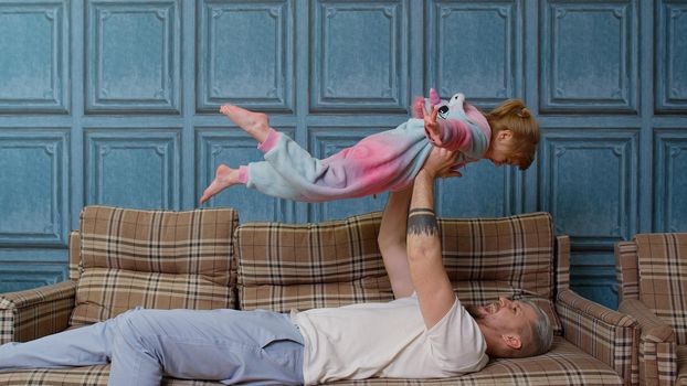 Joyful child girl playing plane game laughing having fun with young father, happy dad lifting small kid daughter up fly enjoying family leisure airplane game relaxing on sofa at home in living room