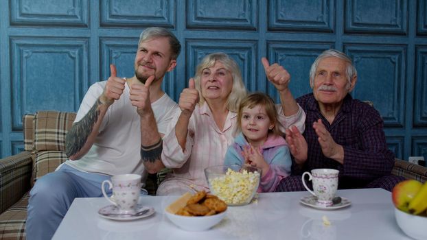 Multigenerational happy family laughing, watching comedy cartoons television movies eating popcorn at home. Senior couple grandfather and grandmother relaxing with adult son and girl kid granddaughter
