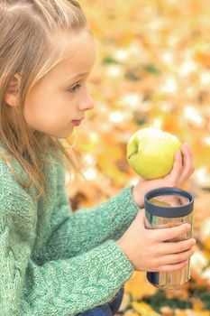 Beautiful little caucasian girl with apple and drink sitting in autumn park