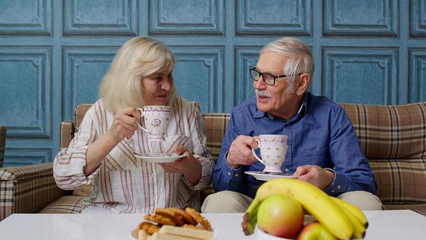 Mature family couple grandfather, grandmother relaxing on cozy sofa, enjoying pleasant conversation, holding cups of hot tea. Loving happy senior aged man talking discussing with elderly smiling wife