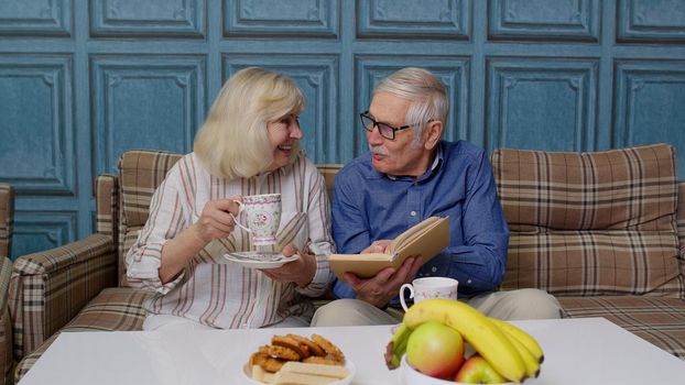 Happy smiling senior couple grandfather, grandmother resting on sofa, drinking coffee, reading book at home. Portrait of cheerful retired husband and wife enjoying tea and talking discussing relaxing