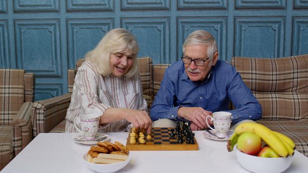 Retired senior couple talking drinking tea, playing chess in modern living room lounge together. Old elderly grandmother, grandfather enjoying coffee having conversation relaxing sit on sofa at home