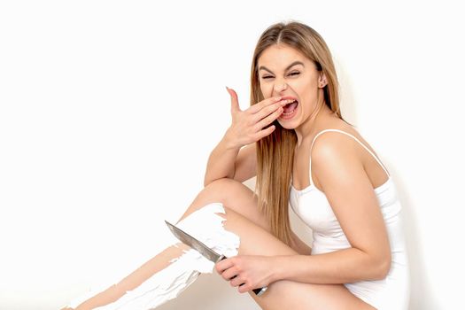 Beautiful young caucasian woman shaves her legs with a knife on a white background