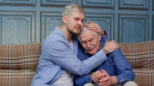 Compassionate young man supporting, giving psychological help to frustrated middle aged father. Grown up son asking for apologizes to offended stressed elderly senior daddy, sitting together on couch