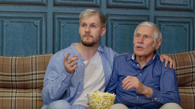 Excited Caucasian family senior man father and adult son sitting on sofa and watching sport game on TV with eating popcorn together. Dad and son cheering soccer or football team to winning the match