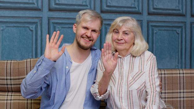 Happy bonding loving generations family of senior mother and handsome adult son or grandson waving hi, hello, welcome. Cheerful excited young man with gray-haired old mature mom or grandmother